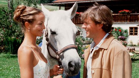 Eine Frau in weißem Kleid und ein Mann schauen sich an. Zwischen ihnen ein weißes Pferd.