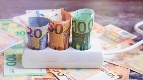 Dreifachsteckdose auf Euro Banknoten. In den einzelnen Steckdosen befinden sich verschiedene eingerollte Geldscheine.