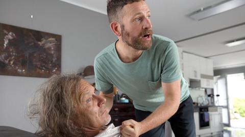 Ein Mann vedroht einen andern Mann auf dessen Sofa.