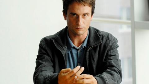 Ein Mann mit schwarzer Jacke schaut streng in die Kamera.