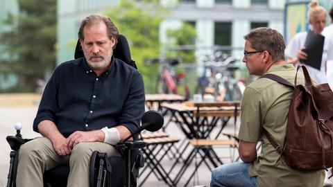 Ein Mann im Rollstuhl, neben ihm sitzt ein Mann mit Brille und Rucksack auf einer Bnak