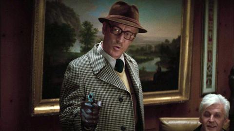 Ein Mann mit Hut und Brille hält eine Pistole in der Hand. Im Hintergrund ein gemaltes Bild.