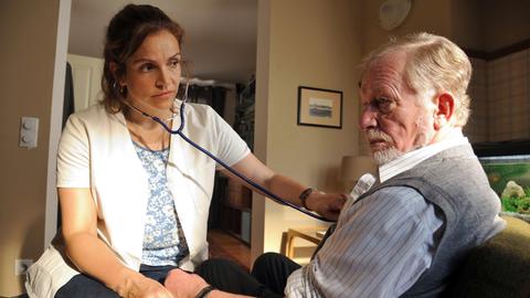 Vera (Rebecca Immanuel) untersucht ihren Patienten (Hermann Beyer) mit einem Stethoskop.