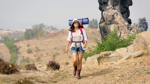 Eine Frau mit Rucksack, Sonnenhut und Sonnnebrille läuft durch die Wüste.
