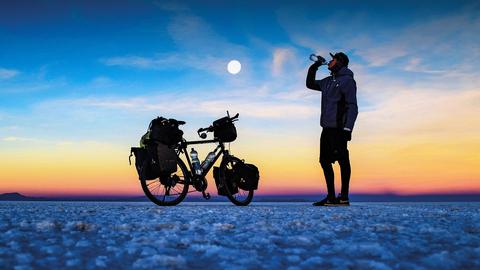 Dennis Kailing steht mit seinem Fahrrad in Boliviens Salar de Uyunineu und trinkt aus einer Wasserflasche. Die Sonne geht unter, der Himmel ist in orange-gelb-blaues Licht getaucht. Hinter ihm strahlt bereits der Mond.