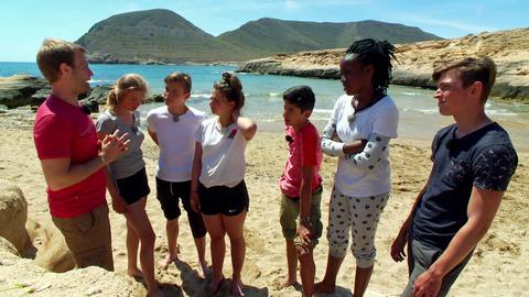 Coach Tobi steht mit den Jugendlichen am Strand und erzählt ihnen etwas.