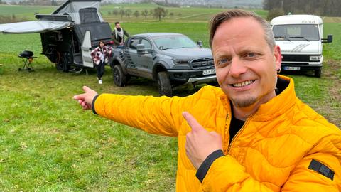 Friso Richter deutet hinter sich auf ein Pärchen und einen Offroad-Wohnwagen.