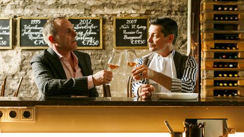 Zwei Männer sitzen an der Bar und stoßen mit einem Glas Wein an.