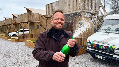 Moderator Friso Richter öffnet eine Sektflasche, im Hintergrund sieht man einen Camper.