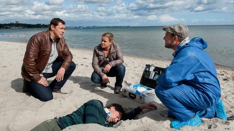Drei Personen knien am Strand um eine Leiche herum.