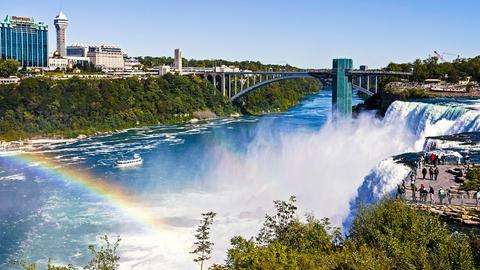 Blick auf die Niagarafälle und die gegenüberliegende Umgebung. Ein Regenbogen erscheint durch das Zusammenspiel des feinen Wassernebels und des sonnigen Wetters.