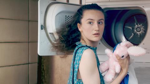 Eine junge Frau steht im Waschsalon vor einer Waschmaschine und hält einen Kuscheltierhasen in der Hand.