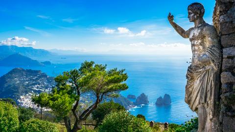 Blick auf Capri vom Mount Solaro aus. Rechts im Bild steht eine lebensgroße steinerne Figur mit nackter Brust und ausgestrecktem Arm.