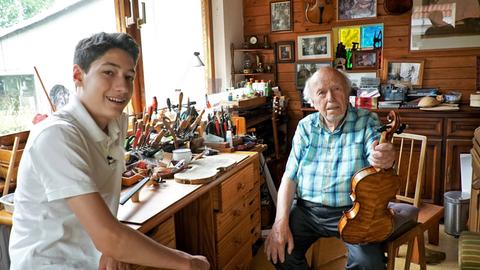 Ein Junge sitzt am Schreibtishc neben einem älteren Mann, der eine Geige in der Hand hält