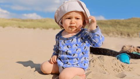 Baby sitzt mit Sonnenhut im Sand, im Hintergrund Dünen und Sandspielzeug.