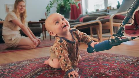 Baby krabbelt auf dem Teppich und greift nach dem Mikrofon des Filmteams, im Hintergrund die Mutter.