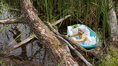 Ein Baby treibt auf einem Körbchen in einem Fluss.